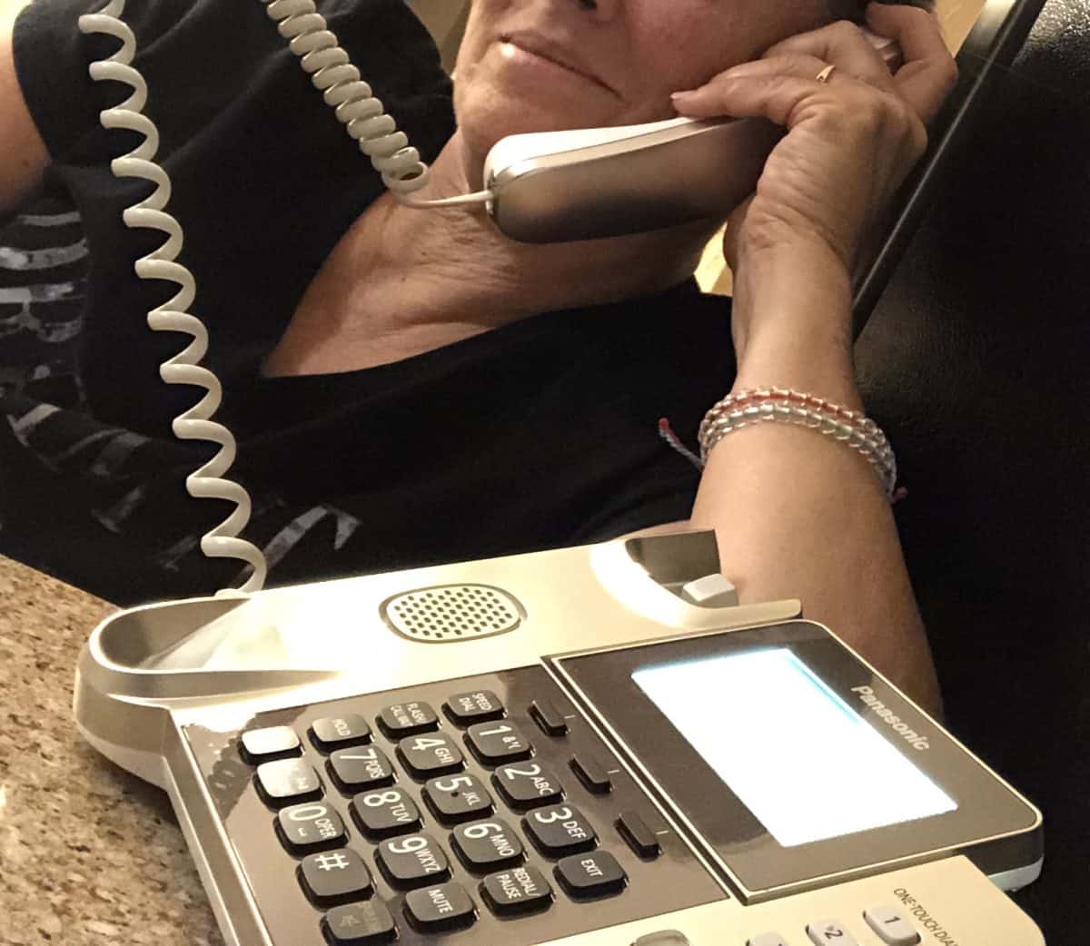 Old Lady Dodge on a push button phone holding up the earpiece and cord in her hand as she is leaning on the table.